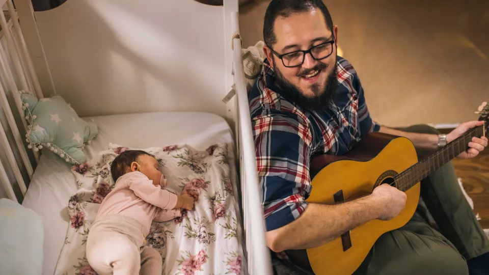 Man with beard and glasses plays guitar beside baby sleeping in crib