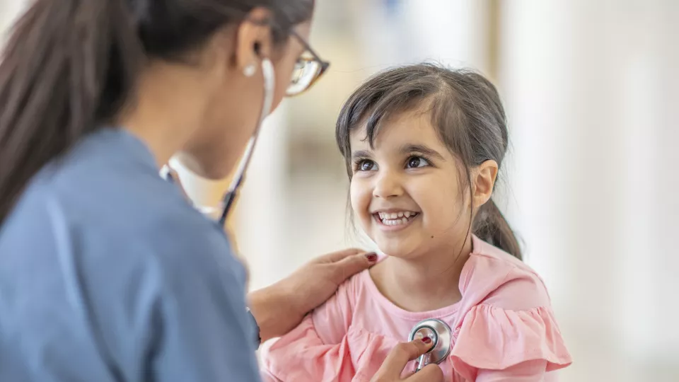 Little girl with medium skin tone smiles as a female health care provider in blue scrubs places a stethoscope on her heart. The girl is wearing a pink top with ruffles.