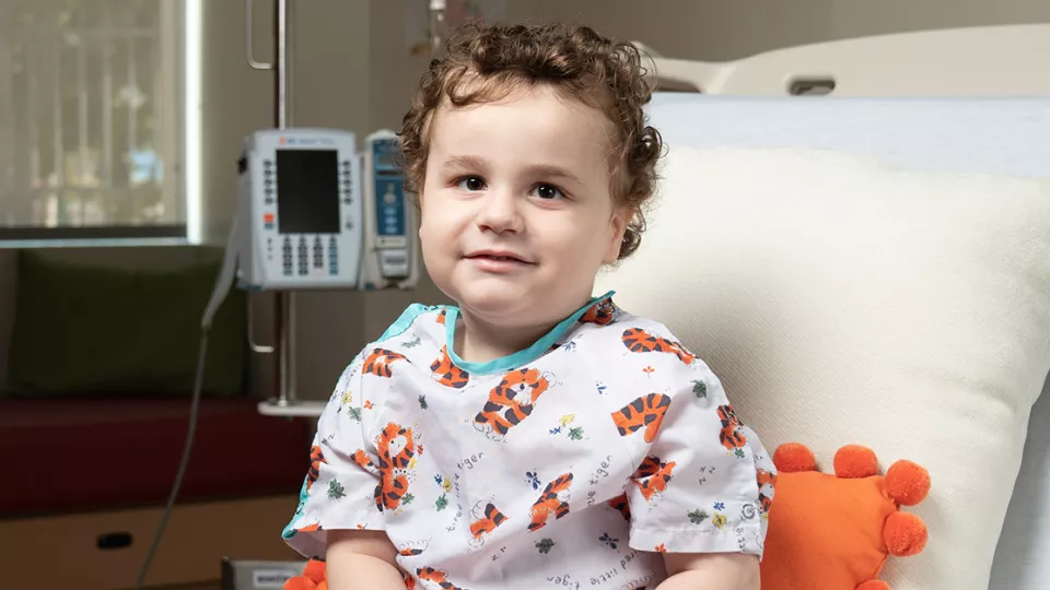 A toddler wearing a medical gown sits on a hospital bed.