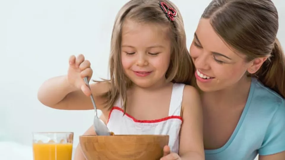 A smiling toddler scoops food from a bowl with a spoon while sitting on their mother's lap.