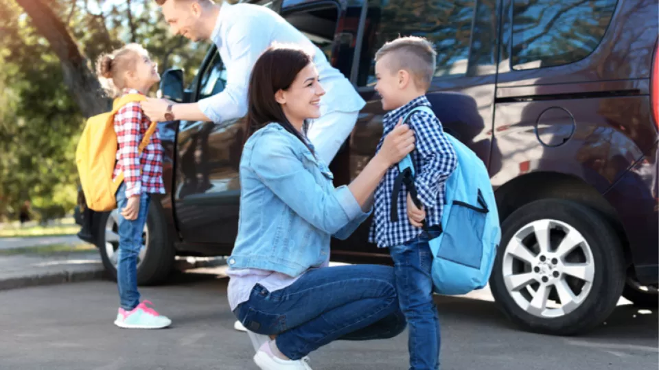 Father and mother with light skin tone smile as they adjust their small children's backpacks