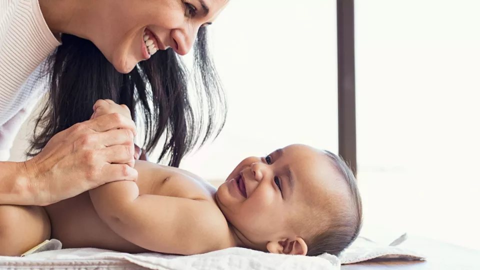 Mother with medium skin tone smiles as she holds the arm of smiling baby with medium skin tone