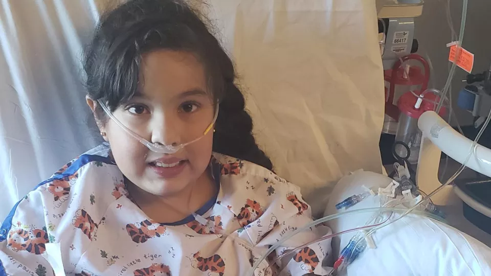 Young girl wearing a medical gown laying in a hospital bed with oxygen tubes at her nose.