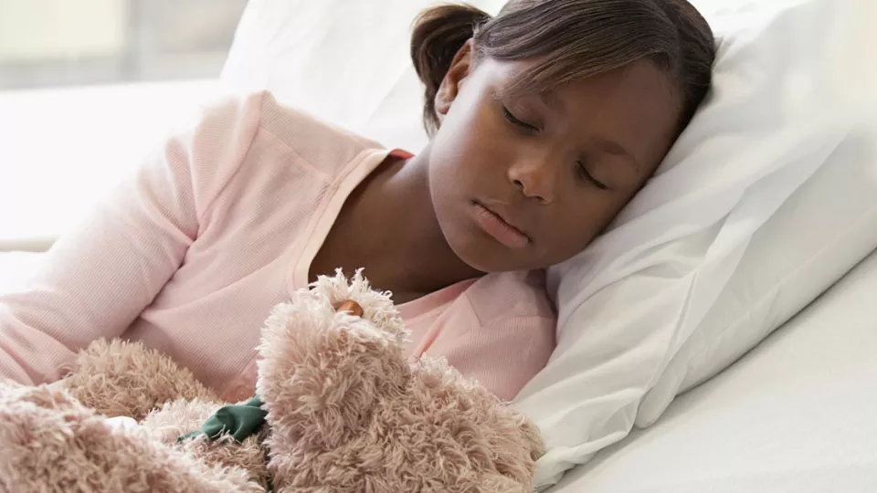 Young girl with dark skin tone and dark hair sleeping with her head resting on her pillow and teddy bear in the foreground