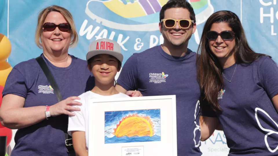 A child holds a painting they made of a sun and sky, while three adults wearing sunglasses pose next to them.