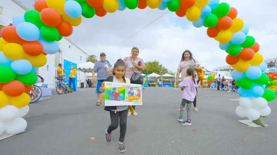 A child holds a Sesame Street toy box while standing under a decorative arch of rainbow balloons.