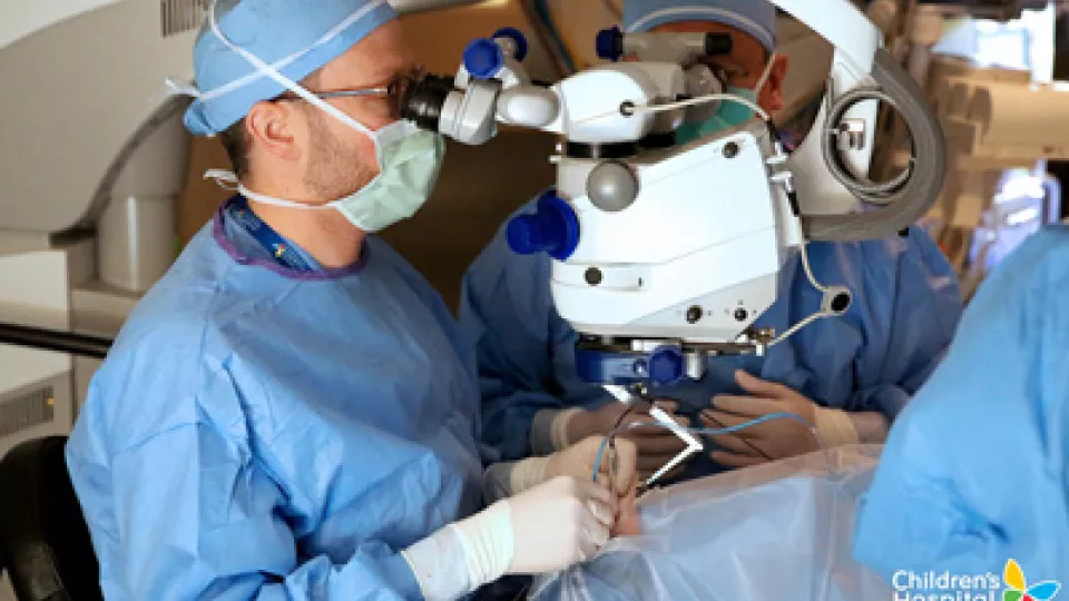 Two men in surgical gowns, head coverings and masks look through a device while performing a procedure