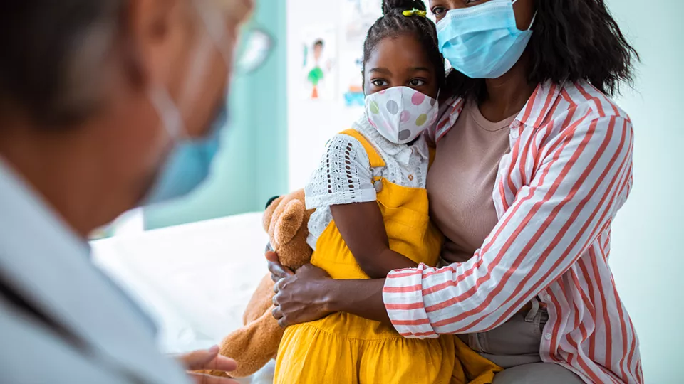 Woman wearing a blue medical procedure, holding a young girl, who is also wearing a medical procedure mask, while talking to a doctor.