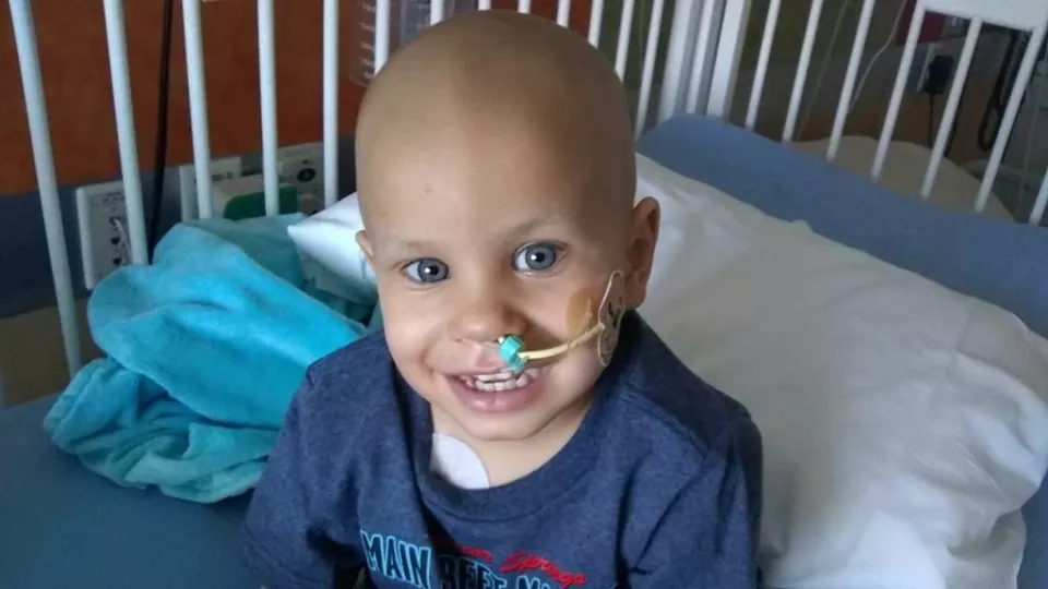 Young child with a bald head and oxygen tube to their nose, wearing a blue shirt sitting in a crib.