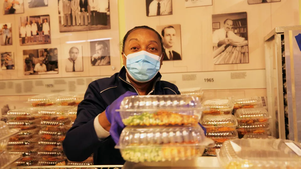 Masked woman with dark skin and wearing surgical mask prepares boxed meals