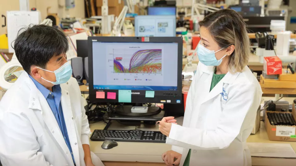 Two masked researchers wearing white lab coats in front of computer monitor in laboratory setting