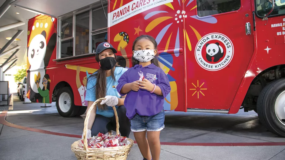 A little girl and a Panda Express worker wear medical procedure masks while nealing next to a Panda Express food truck.