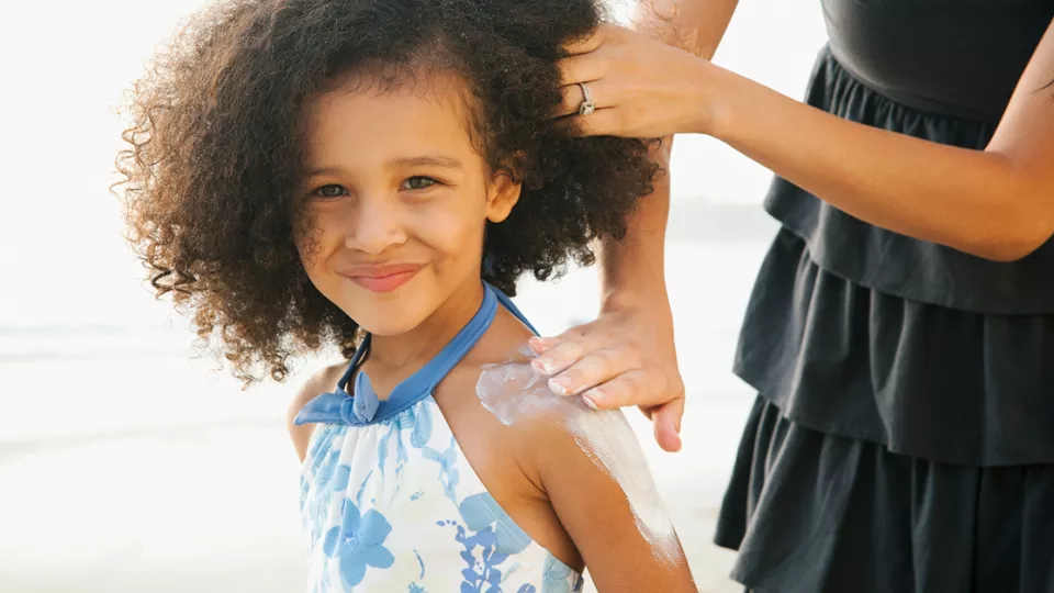 A woman applies sunscreen to the shoulder of a smiling girl with curly hair.