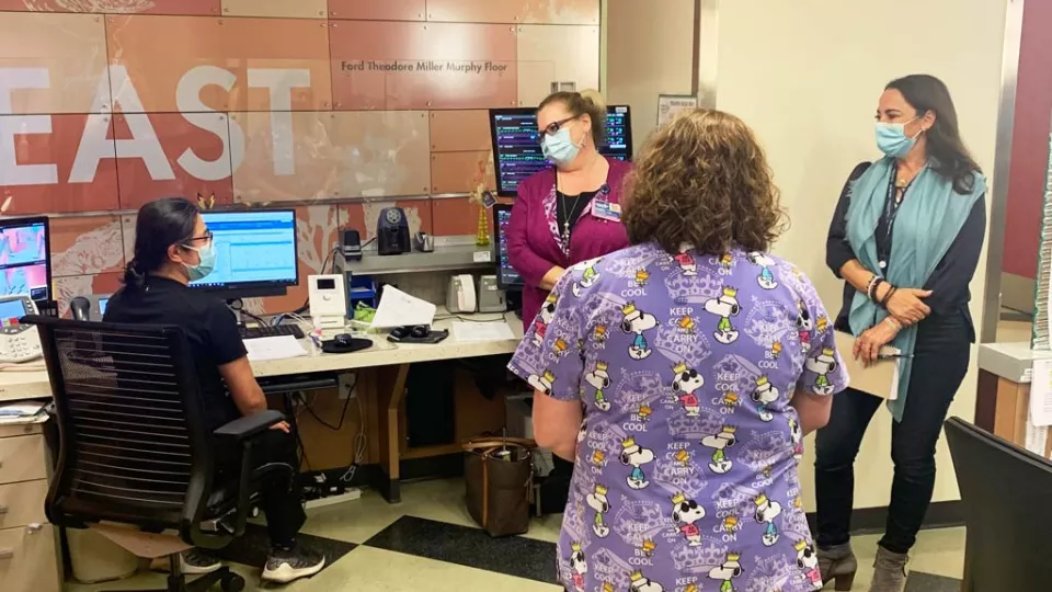 A medium-light skin-toned woman sits at a computer screen, talking with a light skin-toned woman and a medium-light skin-toned woman who stand. All wear surgical masks. A woman in Snoopy scrubs has her back to the camera. 