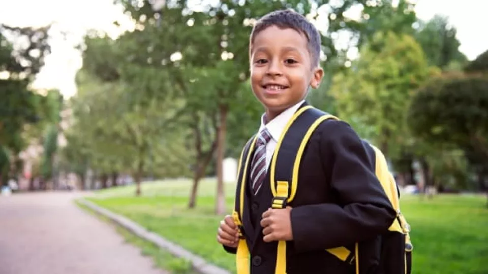 Outdoor image of young boy with medium skin tone and wearing a school uniform and backpack 