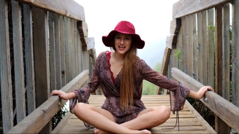 Young woman with light skin tone wearing red beach hat and paisley beach top sits cross-legged on wooden stairs with ocean in the background