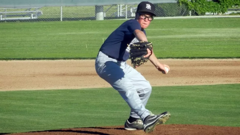 Boy with light skin tone wearing baseball uniform winds up to throw a pitch