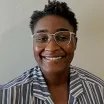 Headshot of a smiling woman with dark skin tone and short dark hair wearing glasses and a blue striped blouse against a neutral indoor background