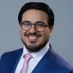 Headshot of a smiling man with medium skin tone, dark hair and close-cropped beard wearing eyeglasses, a white dress shirt and pink tie under a blue suit jacket against a neutral indoor background