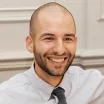 Headshot of a smiling man with medium skin tone, shaved head and a trimmed dark beard wearing a light grey dress shirt and dark grey tie against an indoor background