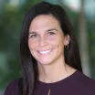 Headshot of woman with light skin tone and dark hair wearing business attire with a blurred outdoor background