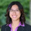 Headshot of a young woman with medium skin tone and dark hair wearing glasses, purple shirt and dark suit jacket with blurred outdoor background