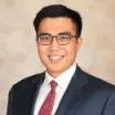 Headshot of man with light skin tone and dark hair wearing rectangular eyeglasses, white dress shirt and red tie under a dark suit jacket against a neutral indoor background