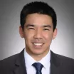 Headshot of a smiling man with medium skin tone and dark hair wearing white shirt with dark tie and suit jacket against neutral background