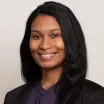 Headshot of a smiling woman with dark skin tone and dark hair wearing purple blouse and dark suit jacket against neutral background