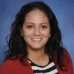 Headshot of a smiling woman with medium skin tone and dark, shoulder-length hair wearing a black and white striped top under a red suit jacket against a neutral blue background