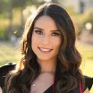 Headshot of a smiling young woman with light skin tone and long brown hair wearing a black top against a blurred outdoor background