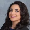 Headshot of a smiling woman with medium skin tone and wavy dark hair wearing a dark suit jacket against a neutral indoor background