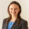 Headshot of a smiling young woman with light skin tone and straight brown hair wearing a light blue top under a dark suit jacket against a neutral indoor background