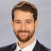 Headshot of a smiling man with medium skin tone, short brown hair and a closely cropped beard wearing a white dress shirt under a dark suit jacket against a neutral indoor background