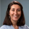 Headshot of a smiling woman with medium skin tone and shoulder-length brown hair wearing a blue suit jacket against a neutral indoor background