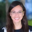 Headshot of a smiling woman with light skin tone and long dark hair wearing eyeglasses, small gold hoop earrings and dark blouse with white floral print against a blurred outdoor background