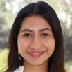 Headshot of a smiling young woman with medium skin tone and shoulder-length brown hair wearing a white blouse against a blurred outdoor background