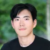 Headshot of a man with light skin tone and short dark hair wearing a black t-shirt against a blurred green outdoor background