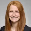 Headshot of a smiling woman with light skin tone and straight auburn hair wearing a dark suit jacket against a neutral indoor background