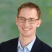 Headshot of a smiling man with light skin tone and auburn hair wearing rectangular eyeglasses and an open-collar light blue dress shirt under a dark suit jacket against a blurred outdoor background