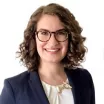 Headshot of a smiling woman with light skin tone and short wavy brown hair wearing tortoise shell glasses and a white blouse under a dark suit jacket against a white background