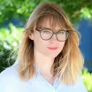 Headshot of a smiling woman with shoulder-length light brown hair wearing large round eyeglasses and a light-colored blouse against a blurred outdoor background