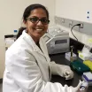 Headshot of a smiling young woman with medium skin tone and dark pulled-back hair wearing rectangular eyeglasses and a white lab coat and gloves in a laboratory setting