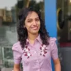 Portrait of a smiling woman with medium skin tone and wavy, shoulder-length dark hair wearing a short-sleeved purple blouse with floral pattern against blurred outdoor background