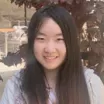 Headshot of a smiling young woman with light skin tone and long straight dark hair wearing a grey hoodie against dark foliage