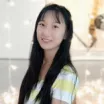 Headshot of a smiling young woman with light skin tone and long straight dark hair wearing a white and yellow striped t-shirt against a bright outdoor background