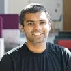 Headshot of a smiling man with dark skin tone and short dark hair wearing a black t-shirt against a blurred indoor background