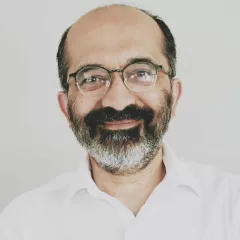 Headshot of a smiling man with light skin tone and receding dark hair wearing rounded glasses and an open-collared white shirt against a neutral background