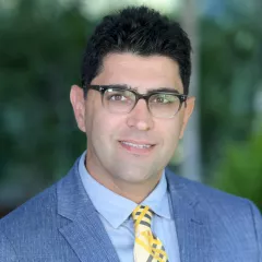 Headshot of a smiling man with light skin tone and dark hair wearing black rimmed glasses, yellow tie and light blue dress shirt under a blue suit jacket against a blurred outdoor background