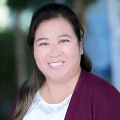 Headshot of a smiling woman with light skin tone and dark hair wearing a purple cardigan against a blurred outdoor background